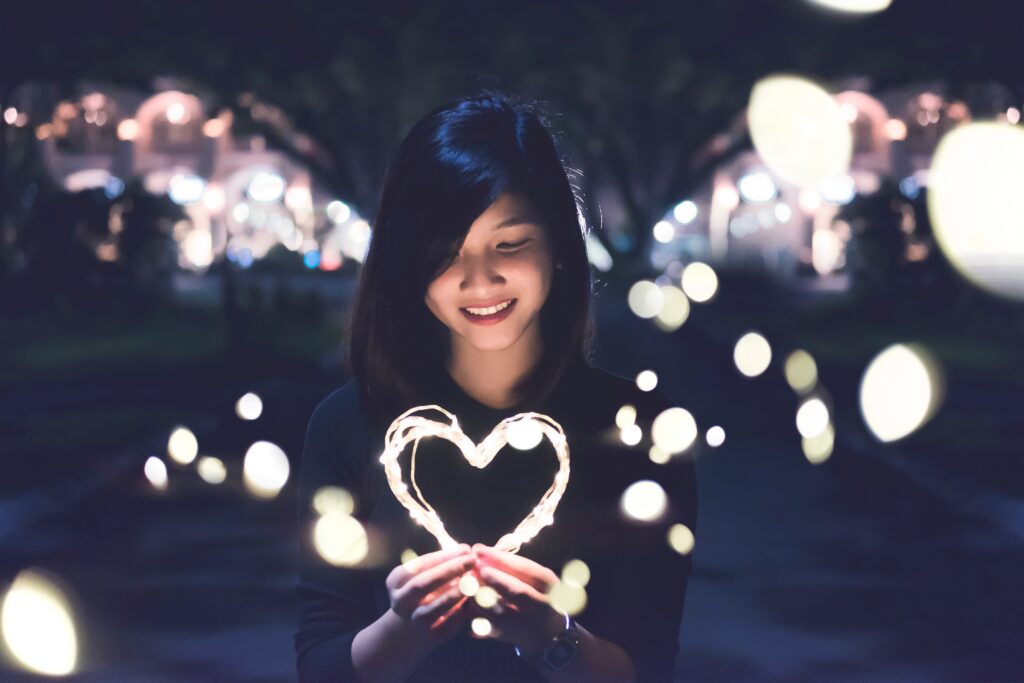 person holding lights that have been formed into a heart shape at night with lights in the background as a visual demonstration of practicing self care